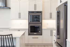 Kitchen with multiple ovens, freestanding refrigerator, light stone countertops, white cabinetry, and light wood finished floors