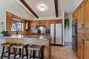 Kitchen featuring wood finish cabinetry, dark stone counters, a peninsula, freestanding refrigerator, and vaulted ceiling with beams