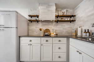 Indoor bar with open shelves, white cabinetry, freestanding refrigerator, backsplash, and dark stone countertops
