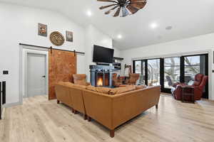 Great room featuring a warm lit fireplace, a ceiling fan, a barn door, light wood-style floors, and vaulted ceiling