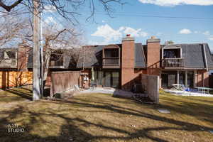 Back of house with a balcony, a patio area, a chimney, a lawn, and a shingled roof