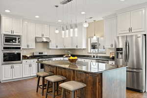 Two tone kitchen with stainless steel appliances, dark stone countertops, pendant lighting, a kitchen bar, and a center island