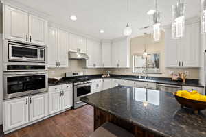 Kitchen featuring dark stone counters, stainless steel appliances, white cabinets, dark wood finished floors, and hanging light fixtures