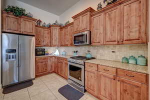 Kitchen featuring stainless steel appliances, light stone countertops, light tile patterned flooring, wood finish cabinetry, and backsplash