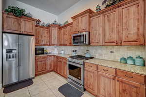 Kitchen with stainless steel appliances, light stone countertops, light tile patterned floors, wood finish cabinetry, and decorative backsplash