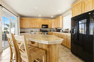 Kitchen featuring black appliances, light wood finish cabinetry, light tile patterned floors, recessed lighting, and a kitchen breakfast bar