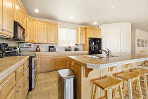 Kitchen featuring black appliances, light wood finish cabinetry, an island with sink, light tile patterned floors, and a kitchen bar