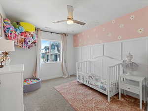 Bedroom with wallpapered walls, light colored carpet, ceiling fan, wainscoting, and a nursery area