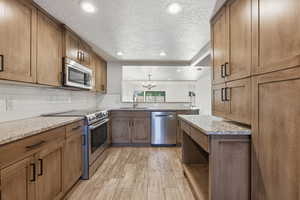 Kitchen featuring stainless steel appliances, wood finish cabinetry, light stone countertops, light wood-style flooring, and a textured ceiling