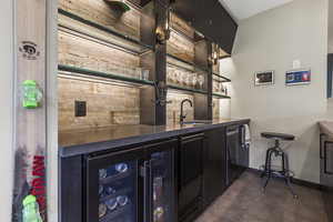 Indoor wet bar with open shelves, wine cooler, dark cabinetry, dark countertops, and dishwashing machine