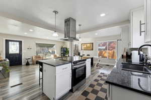 Open floor plan Kitchen with white cabinetry, stainless steel electric range oven, dark stone countertops, a kitchen breakfast bar, and island exhaust hood