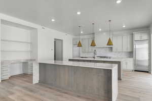 Dual tone kitchen with light stone countertops, light wood-style floors, hanging light fixtures, stainless steel built in refrigerator, and two tone cabinets