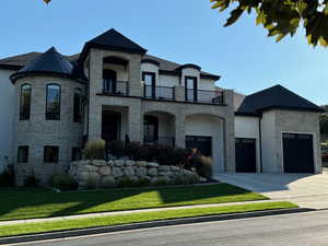 French country home featuring stone siding, stucco siding, a balcony, concrete driveway, and a front yard