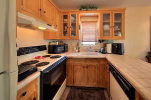 Apartment kitchen with white appliances, tile counters, wood finish cabinets, a peninsula, and glass fronted cabinets