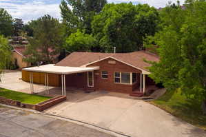 Single story home with brick siding, covered porch, a front lawn, a shingled roof, and view of wooded area