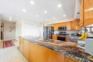Kitchen with dark stone counters, stainless steel appliances, light carpet, wood finish cabinetry, and a peninsula