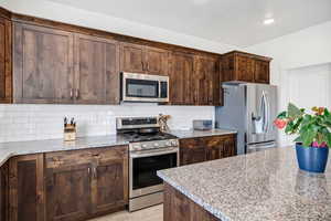 Kitchen featuring stainless steel appliances, light stone countertops, dark wood finish cabinets, decorative backsplash, and light wood-type flooring