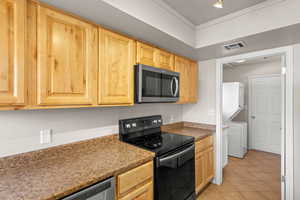 Kitchen with stainless steel appliances, light wood finish cabinetry, dark countertops, light tile patterned floors, and stacked washing machine and dryer