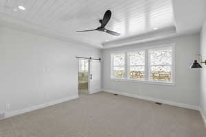 Unfurnished bedroom featuring light carpet, a barn door, a wood tray ceiling, connected bathroom, and a ceiling fan