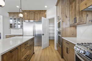 Dual tone kitchen with built in appliances, light wood finished floors, light stone counters, glass fronted cabinets, and decorative light fixtures