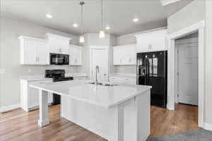 Kitchen featuring black appliances, white cabinets, an island with sink, decorative light fixtures, and light stone counters