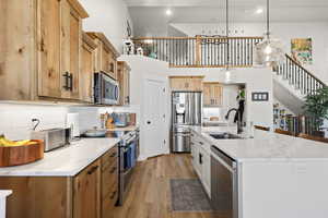 Kitchen with light wood-type flooring, stainless steel appliances, hanging light fixtures, a center island with sink, and light stone counters