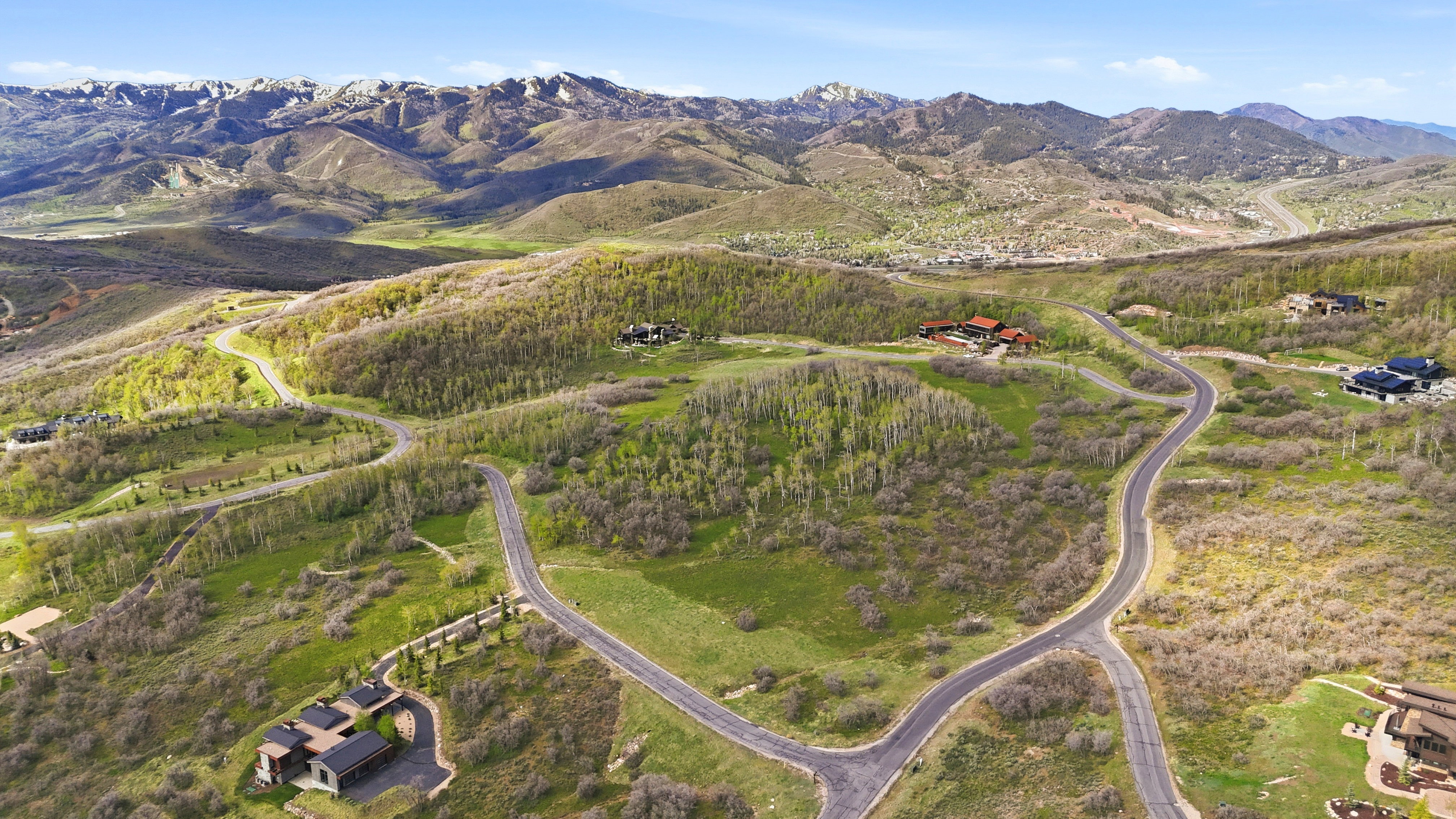 Expansive mountain landscape featuring paved winding roads, scattered residential properties, lush green hillsides, and distant snow-capped peaks