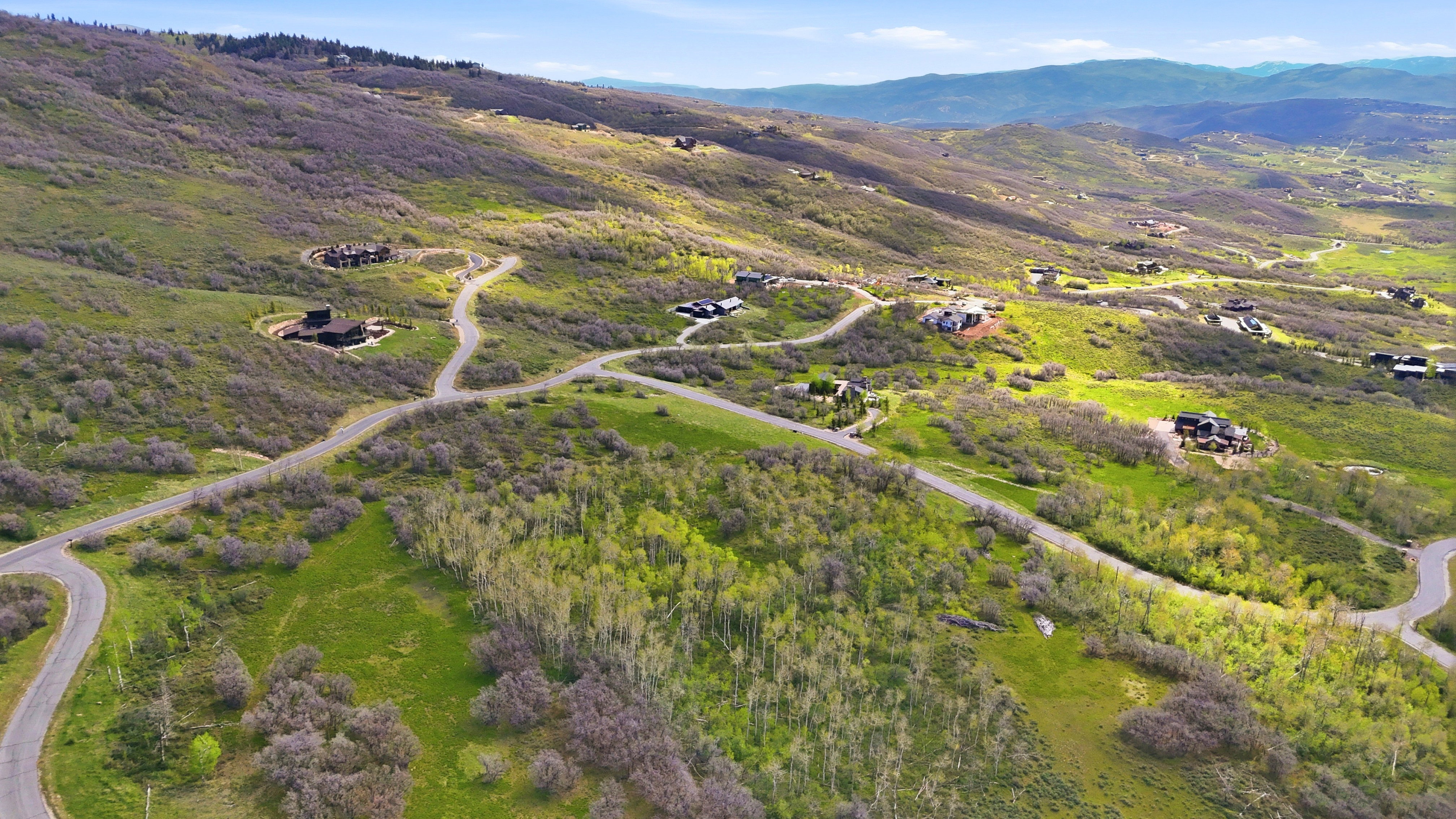 Expansive natural landscape featuring rolling hills, scattered trees, paved roads, and several residential structures nestled within the terrain