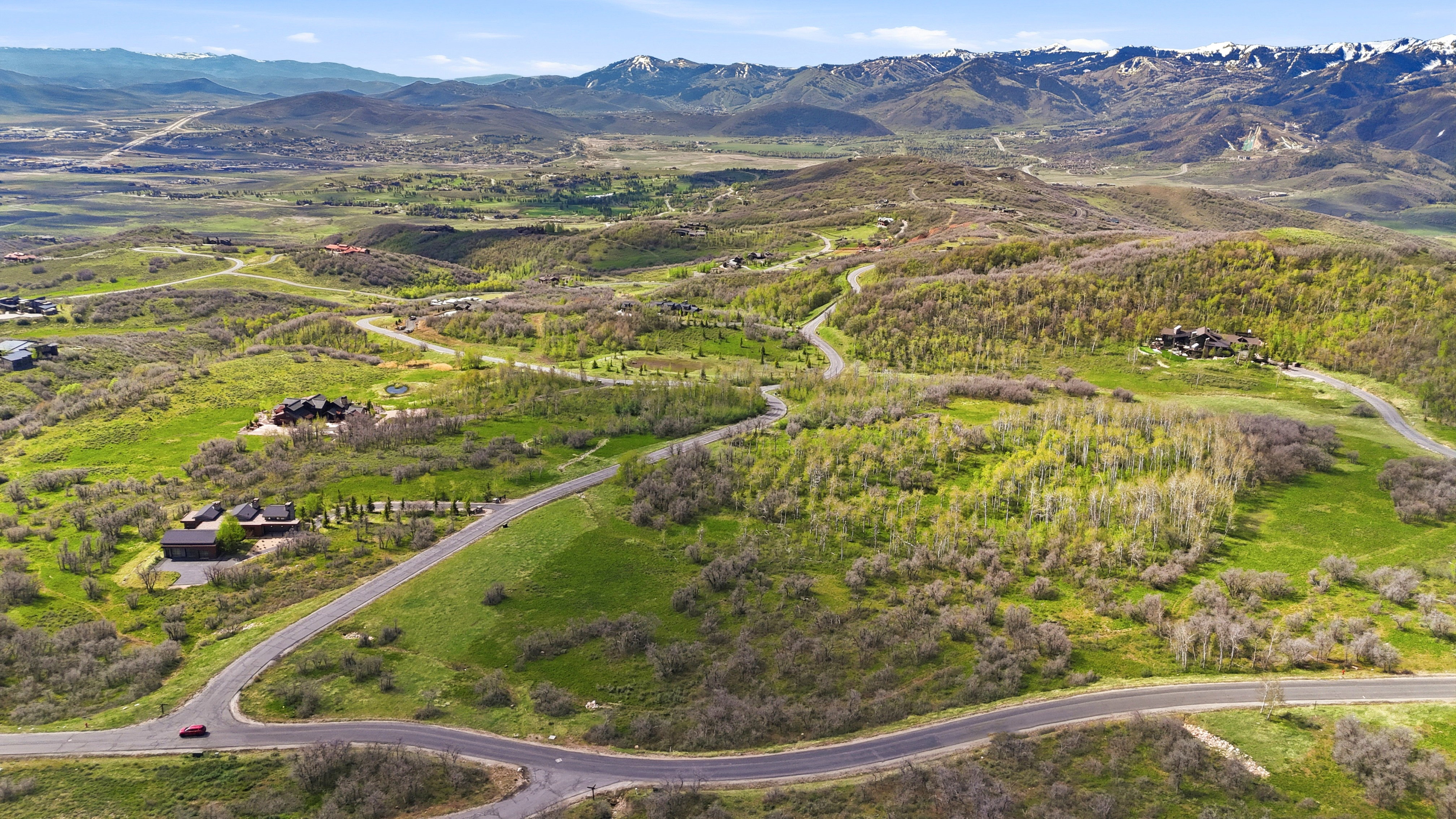 Expansive mountainous landscape featuring winding paved roads, varied topography, lush green foliage, and distant snow-capped peaks
