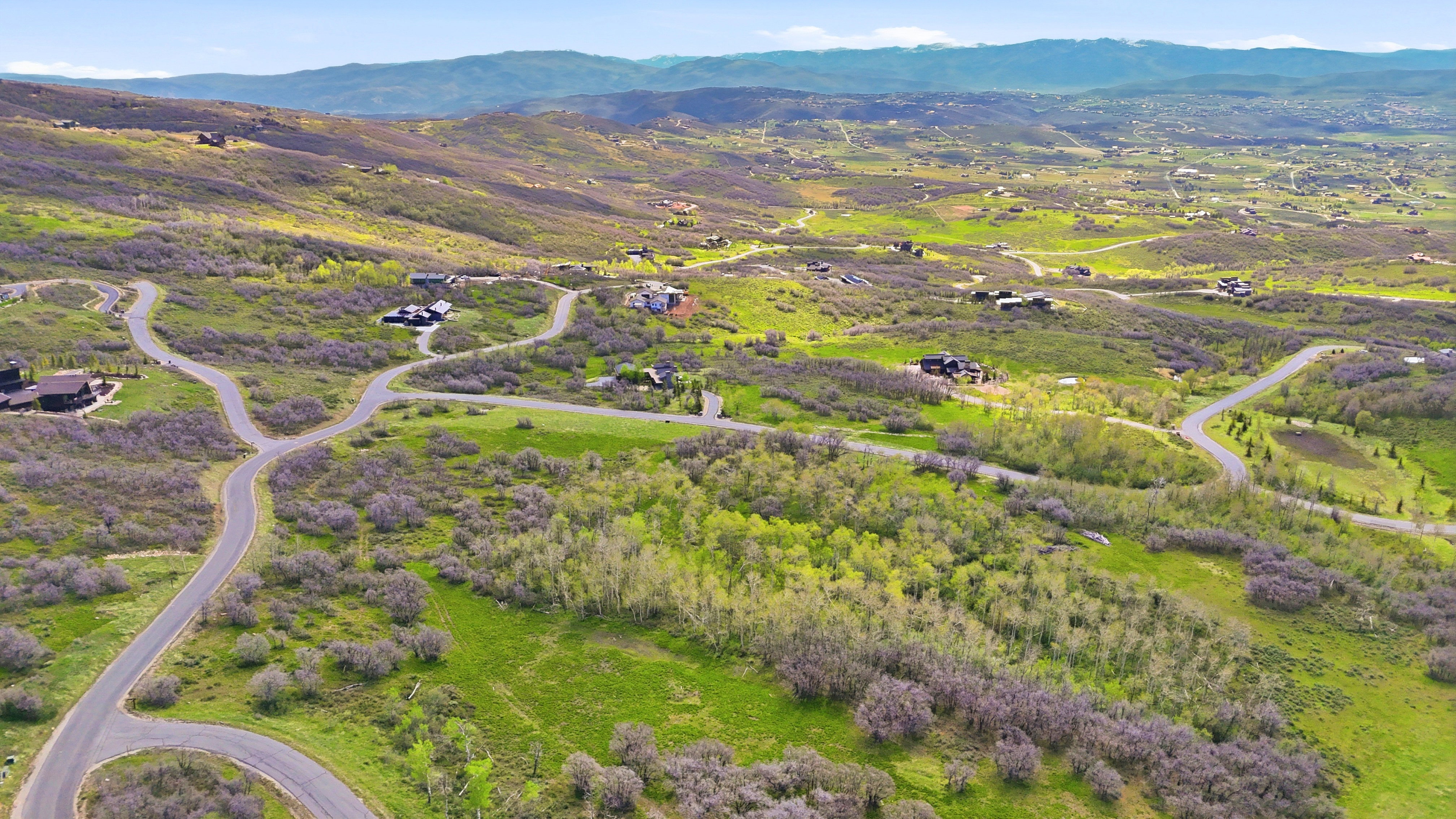 Winding paved roadways traverse a sprawling residential area featuring scattered homes amidst verdant hillsides and distant mountain ranges