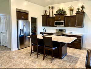 Kitchen with dark wood finish cabinets, stainless steel appliances, a breakfast bar area, light stone countertops, and an island with sink