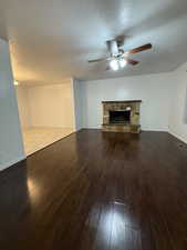Unfurnished living room with a textured ceiling, a stone fireplace, dark wood-style floors, and a ceiling fan