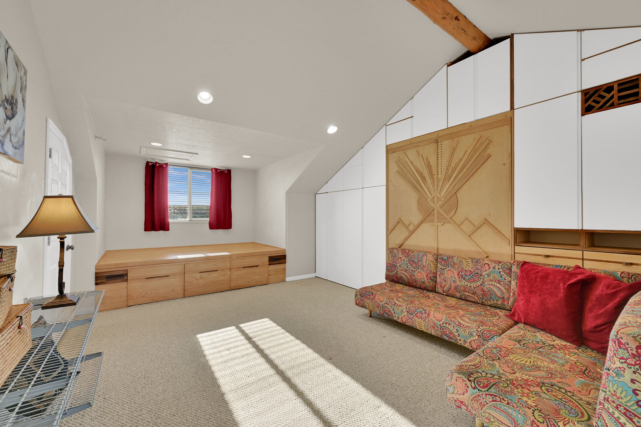 Carpeted room featuring a built-in wooden platform with integrated drawers, recessed lighting, and a window with red drapes