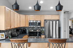 Kitchen with dark countertops, light wood finish cabinets, stainless steel appliances, pendant lighting, and tasteful backsplash