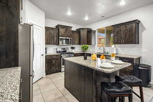 Kitchen featuring dark wood finish cabinetry, stainless steel appliances, light stone countertops, light tile patterned floors, and a center island