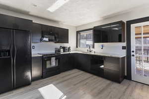 Kitchen with black appliances, dark cabinetry, light stone countertops, light wood-type flooring, and a textured ceiling