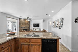 Kitchen featuring a tiled fireplace, black dishwasher, wood finish cabinetry, light stone counters, and a ceiling fan