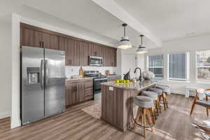 Kitchen with light stone counters, stainless steel appliances, a kitchen island with sink, dark wood-type flooring, and decorative light fixtures