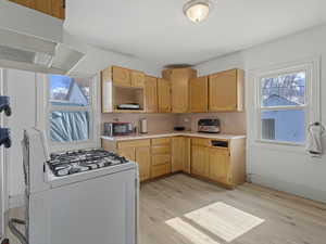 Kitchen with white range with gas stovetop, light countertops, range hood, and light wood finish cabinetry