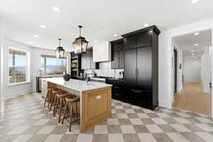 Two tone kitchen with a breakfast bar area, dual tone cabinetry, an island with sink, pendant lighting, and light stone counters