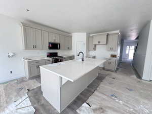 Kitchen featuring gray cabinetry, stainless steel appliances, an island with sink, light stone countertops, and a breakfast bar