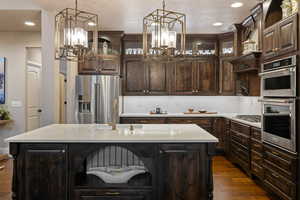 Kitchen with dark wood finish cabinets, stainless steel appliances, a center island with sink, glass fronted cabinets, and dark wood-style flooring