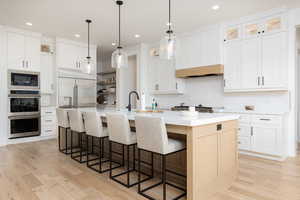 Two tone kitchen with tasteful backsplash, built in appliances, glass fronted cabinets, a kitchen island with sink, and light wood-style floors