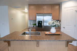 Kitchen with a kitchen bar, stainless steel fridge, wood finish cabinets, a kitchen island with sink, and dark stone countertops