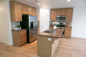 Kitchen featuring stainless steel appliances, a kitchen island with sink, dark countertops, light wood-style floors, and recessed lighting