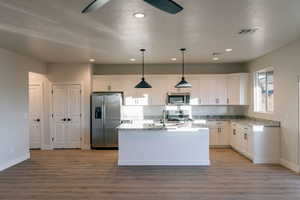 Kitchen with stainless steel appliances, decorative light fixtures, white cabinets, a center island with sink, and light stone counters