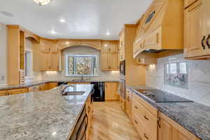 Kitchen featuring light wood finish cabinets, light wood-type flooring, dark stone countertops, black appliances, and tasteful backsplash