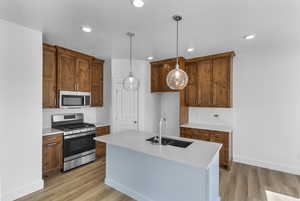 Kitchen with stainless steel appliances, a kitchen island with sink, decorative light fixtures, wood finish cabinetry, and light stone counters