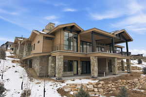 Snow covered back of property with stone siding, a chimney, a patio area, and a balcony