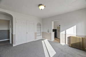 Unfurnished bedroom with radiator, freestanding refrigerator, carpet, arched walkways, and a textured ceiling